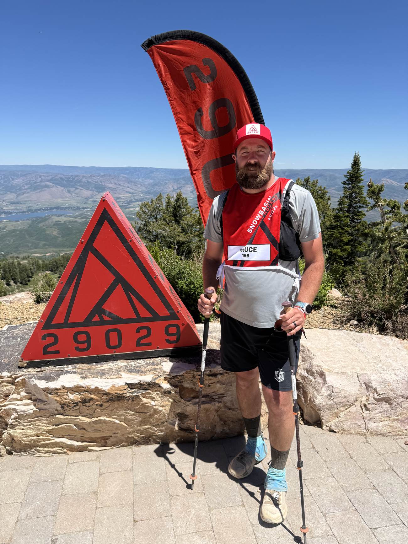 Bruce with trekking poles beside the red 29029 summit marker, the valley far below